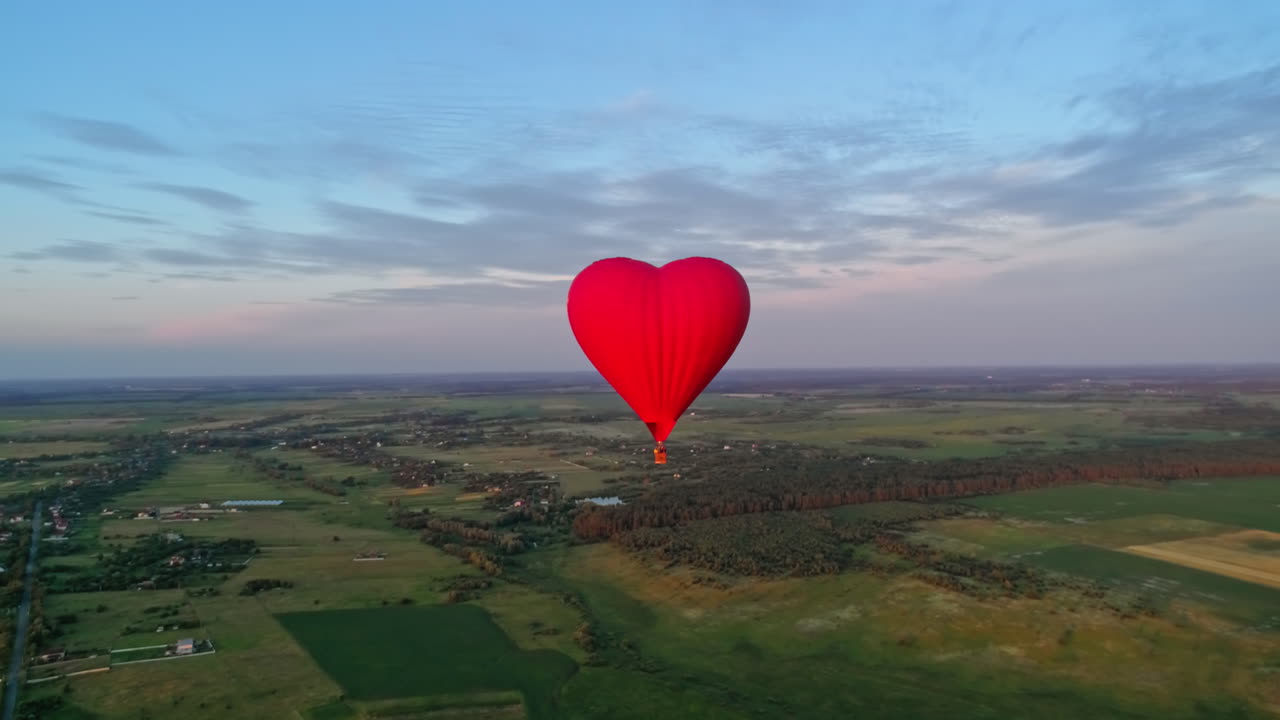 Flight of a red airship. Beautiful hot air balloon in heart shape travelling over the green fields in the morning. Aerostat with basket in the sky.