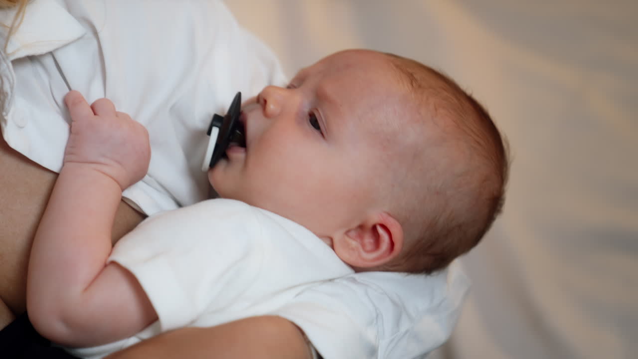 Tiny newborn is waved in mom's hands. Baby boy suckling a pacifier and looking at mom. Close up.