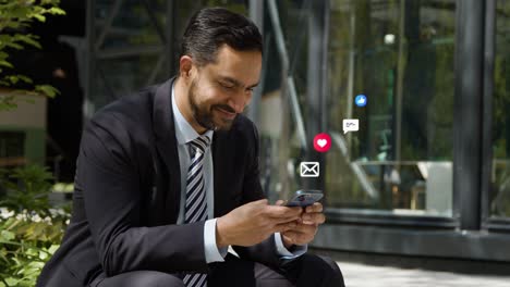 hombre de negocios sentado fuera de las oficinas de la ciudad mirando un teléfono móvil con emojis de gráficos de movimiento que muestran múltiples mensajes y notificaciones de redes sociales