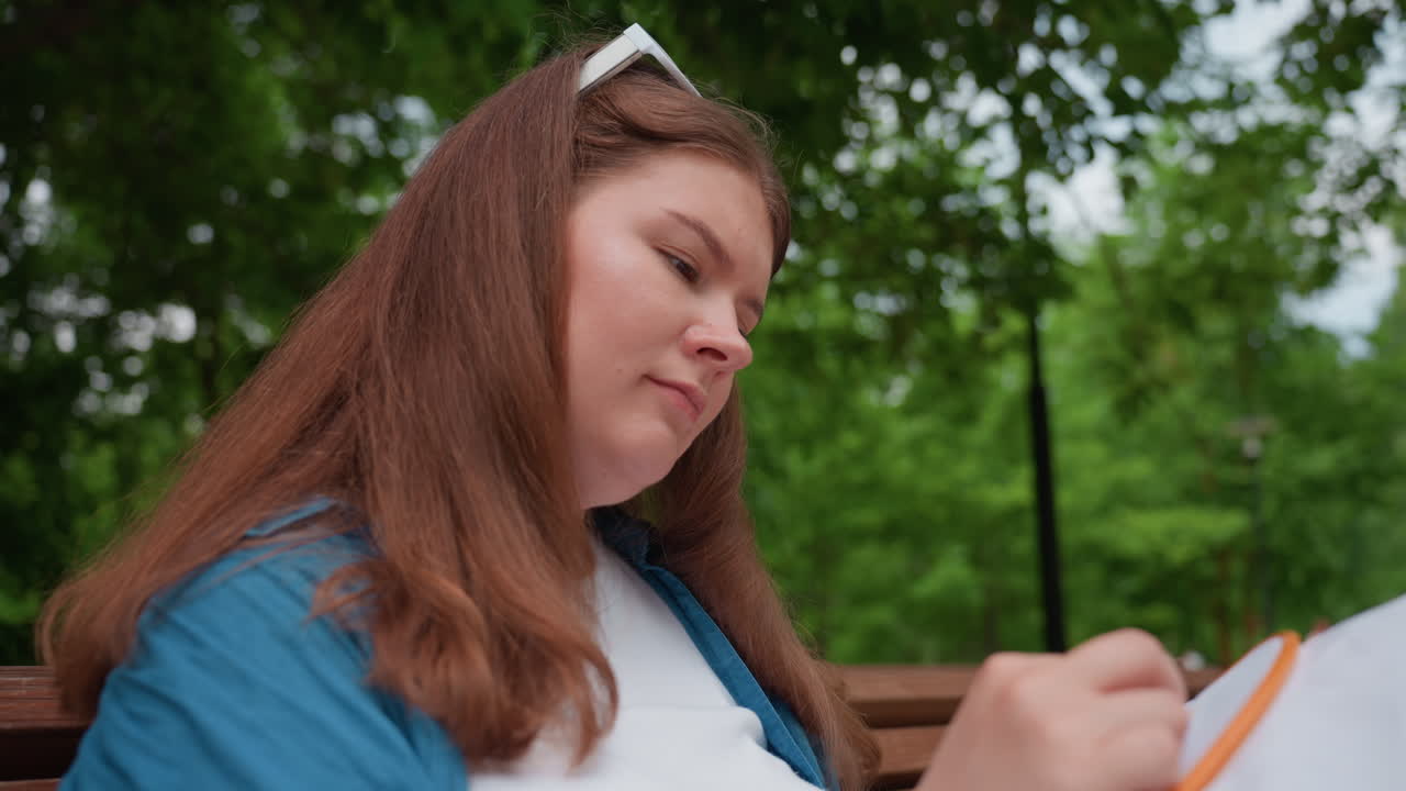 Thoughtful lady sitting on park bench carefully embroidering white fabric with needle, enjoying calm creative process surrounded by green trees, sunlight, and peaceful atmosphere