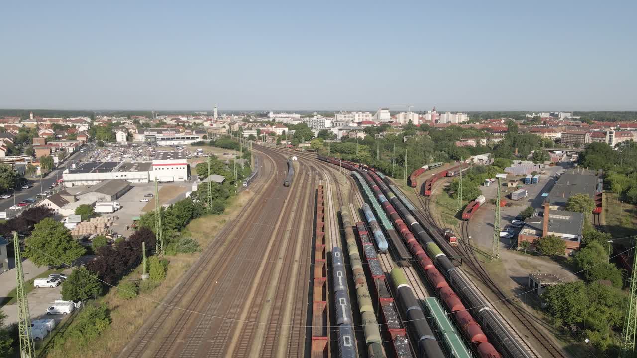 Aerial view of a large railway freight yard adjacent to a small city. Multiple cargo trains and tracks are visible, surrounded by industrial buildings and residential areas under clear skies.