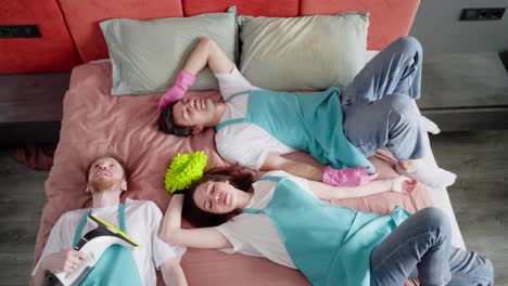 Top-view-of-tired-two-guy-cleaners-in-a-white-T-shirt-and-a-blue-apron-together-with-their-colleague-a-brunette-cleaning-lady-girl-fall-on-a-made-bed-with-a-pink-bedspread-in-a-modern-apartment.-After-ordering-a-cleaning-company