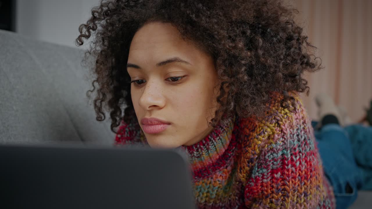 primer plano de una joven afroamericana enfocada navegando por la web en una computadora portátil, acostada en la cama y leyendo noticias en línea, en cámara lenta