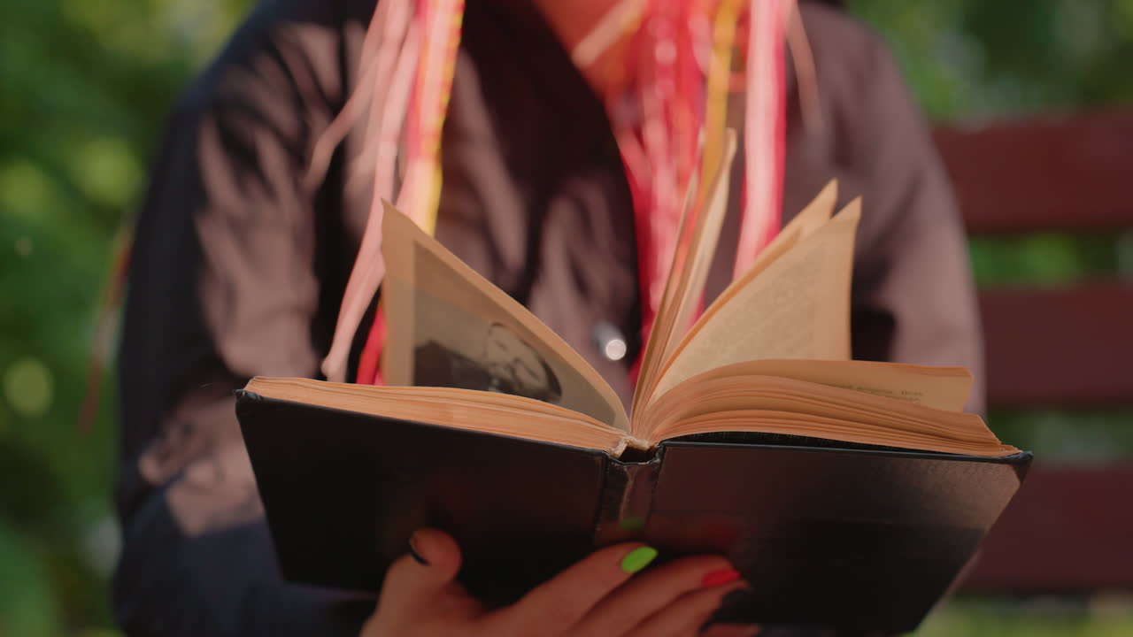 Persona leyendo un libro en un banco a la luz del sol, sintiendo el calor mientras pasa las páginas, con rastas de colores y uñas neón, parque tranquilo de fondo, primer plano de las manos y de un libro de tapa dura abierto, bokeh suave y ambiente reflexivo