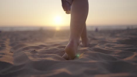 Woman-walks-barefoot-on-sandy-beach-toward-sunset,-slomo-forward-tracking