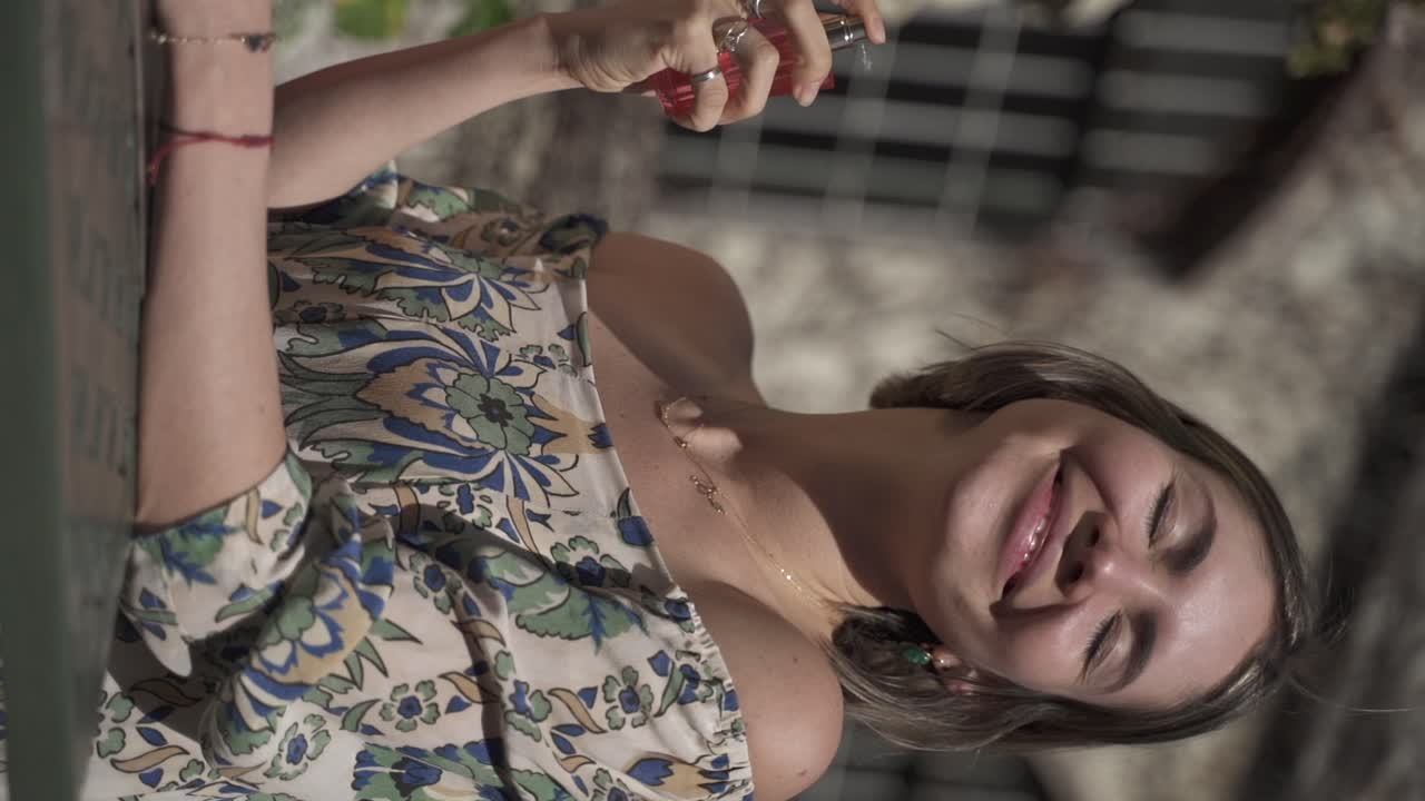 una filmación estacionaria de una mujer alegre sosteniendo una botella de perfume y probándosela en la piel alrededor del cuello