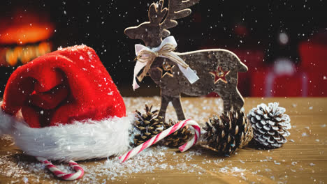 Animation-of-snow-falling-over-pine-cones-candycane,-santa-hat-and-reindeer-on-wood-board