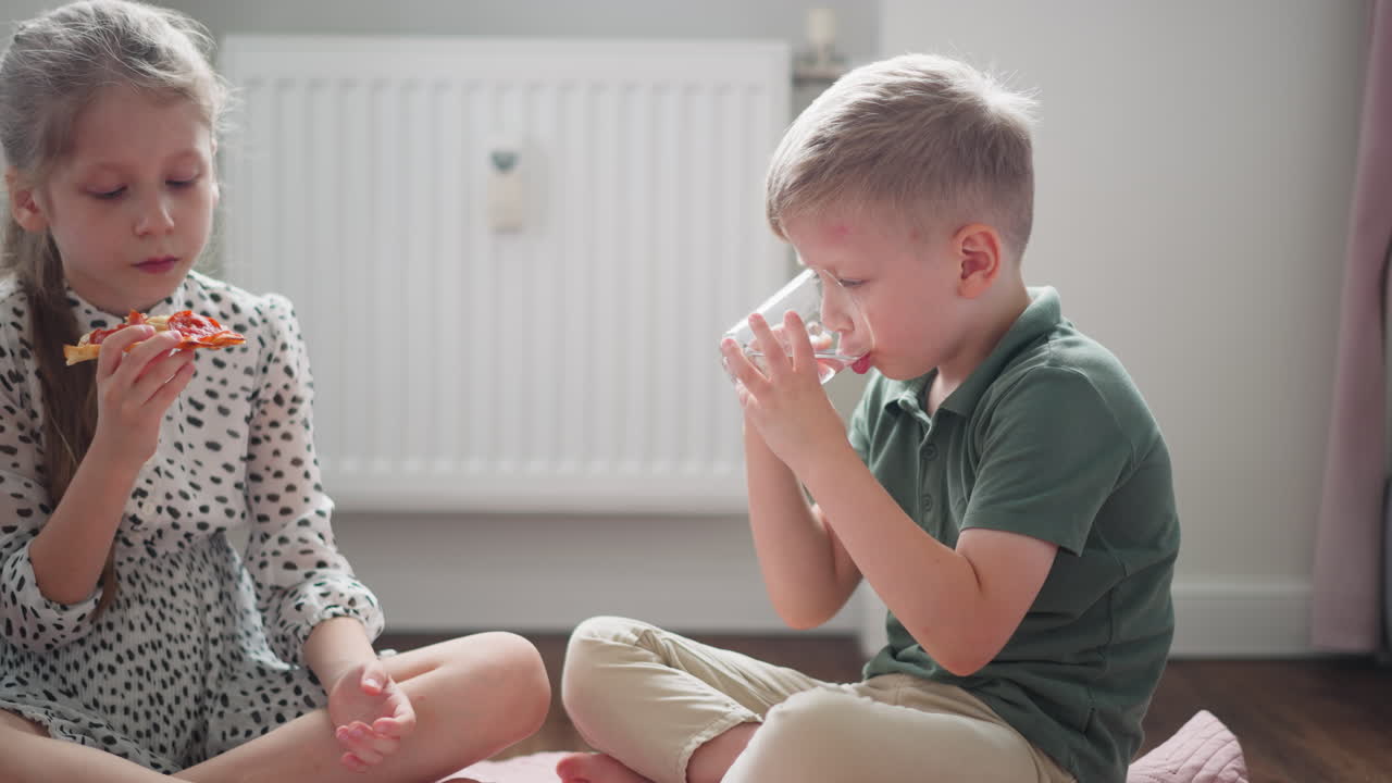 Boy in green shirt drinking water from transparent glass while girl in spotted dress looks at her pizza slice, both seated comfortably indoors on soft mat