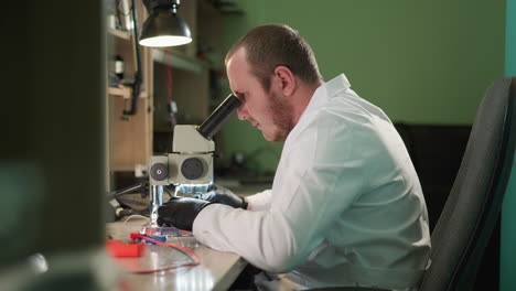 un técnico enfocado en una bata de laboratorio blanca trabaja en un circuito bajo un microscopio en un entorno de laboratorio, el espacio de trabajo está equipado con varios instrumentos para el trabajo, incluida una lámpara de escritorio