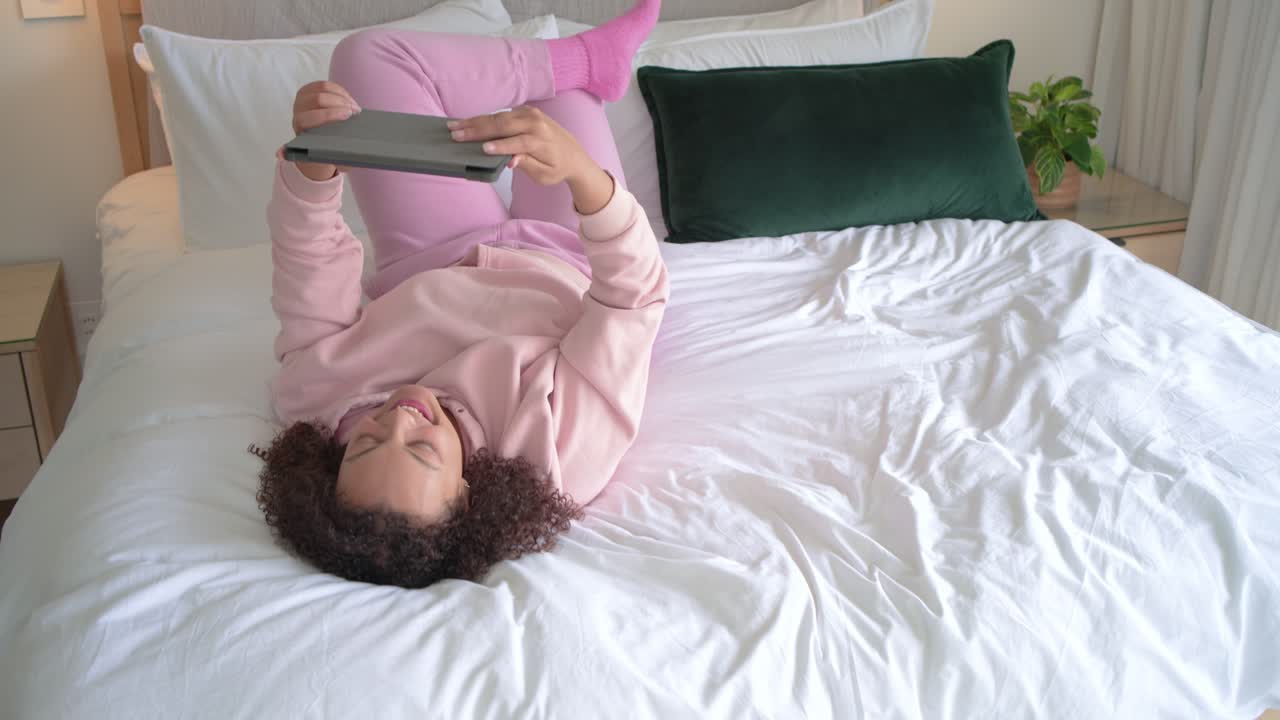 Woman wearing pink hoodie, propping on elbows on bed, reading tablet, reacting and laughing