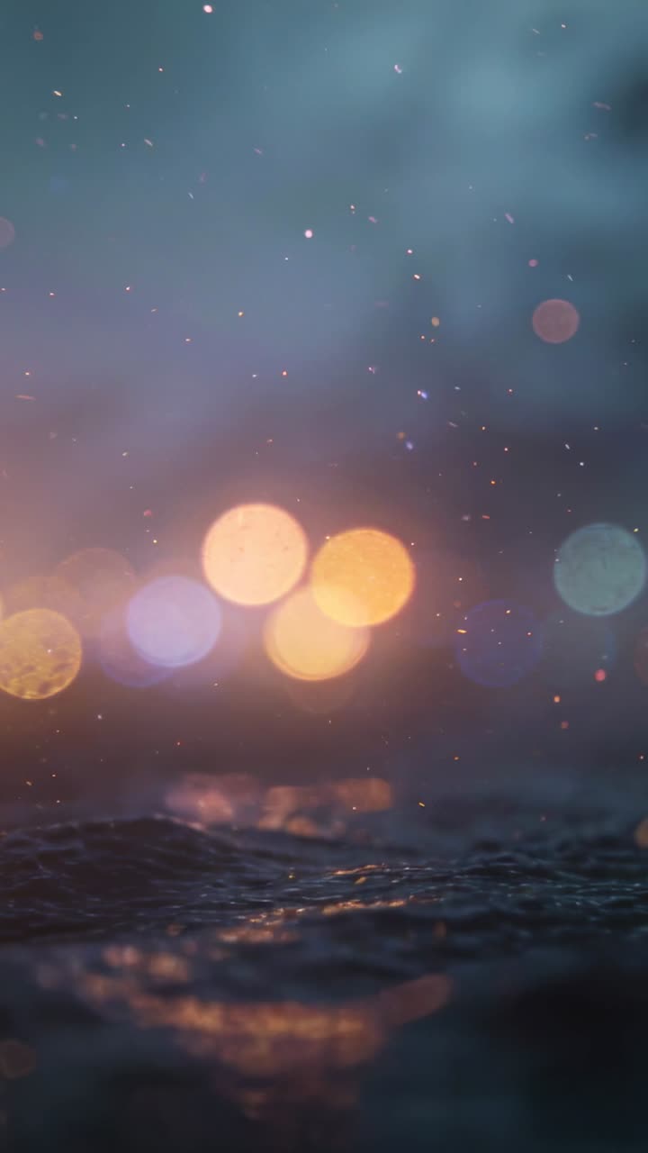 Vertical video: Shifting low cam filming rippling sea at dusk showing warm bokeh lights and spray