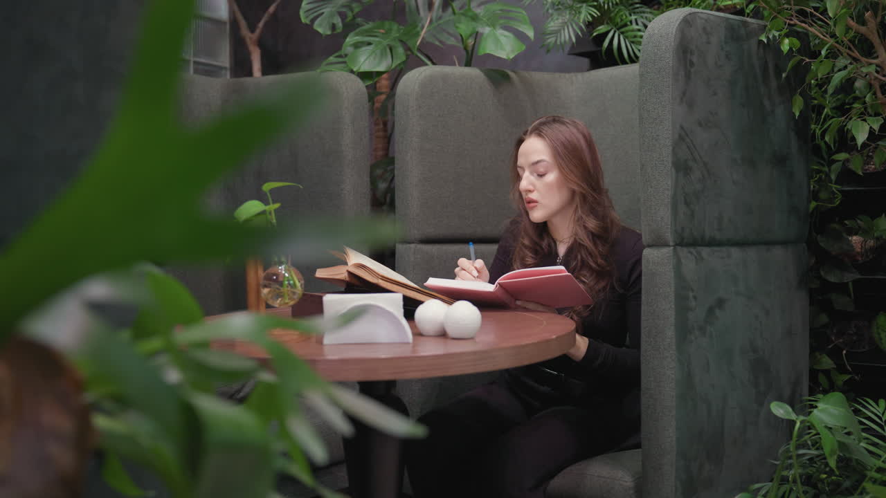 Lady sits inside cozy green lounge booth surrounded by plants, writing in pink book with open brown book nearby on wooden table beside decorative objects and tissue holder