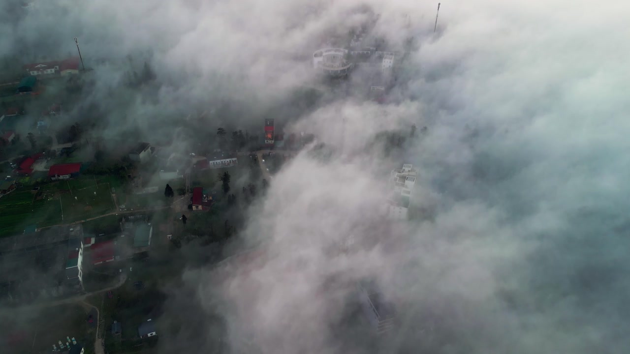 High altitude drone view of a remote highland village covered in thick fog with houses and narrow roads emerging through drifting clouds in the mountainous region of northern Vietnam