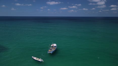 drone moves past a anchored fishing boat and skiff towards a brilliant horizon showing meeting of the turquoise ocean and blue sky