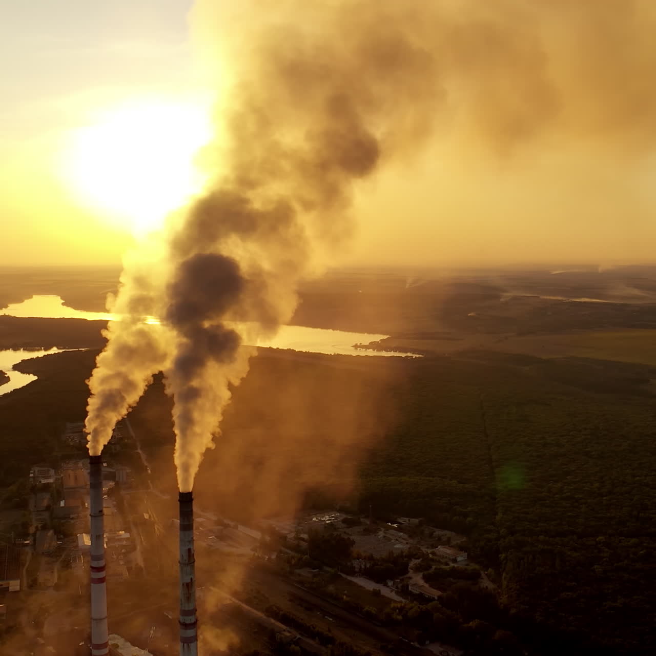 Industrial pipes with smoke in nature. Metallurgical plant among green fields at beautiful sunset. Aerial view. Camera moves right.