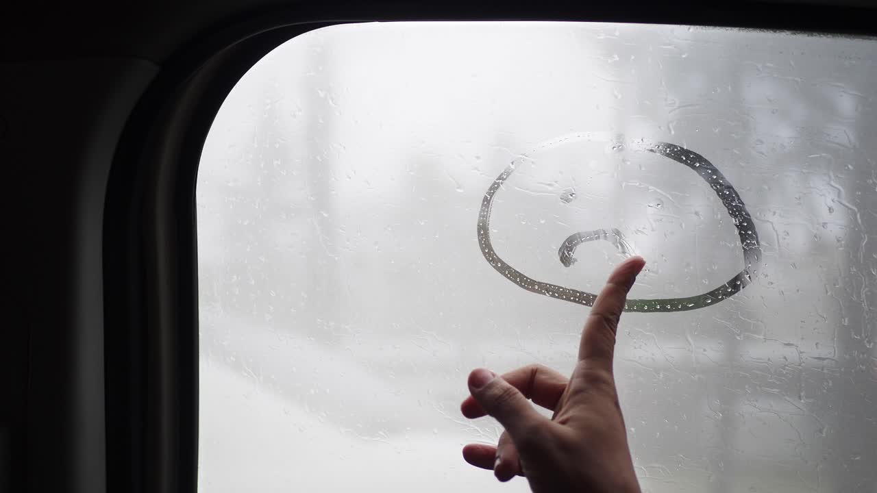 한 손이 비가 오는 차 창문에 슬픈 얼굴을 그린다