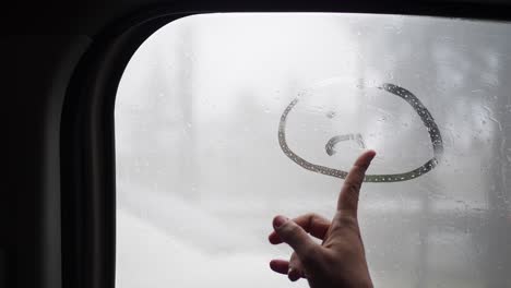 una mano dibuja una cara triste en una ventana de coche lluvioso