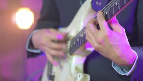 cerca de un músico profesional con traje negro tocando acordes rápidos en una guitarra eléctrica stratocaster durante una sesión en vivo en el escenario con cálidas luces de estudio en el fondo borroso