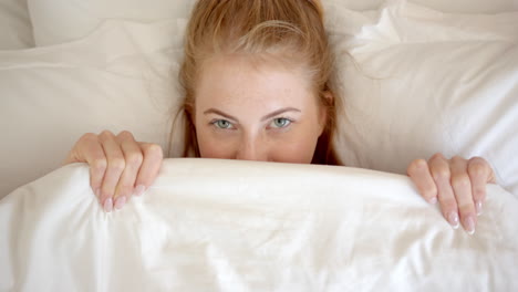 woman peeking from under blanket, relaxing in bed at home