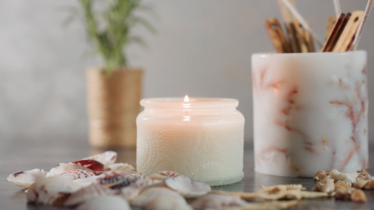 Close up of hand lighting white candle with matchstick, flame catching wick surrounded by seashells, starfish, snail shells on dark marble table with soft natural lighting