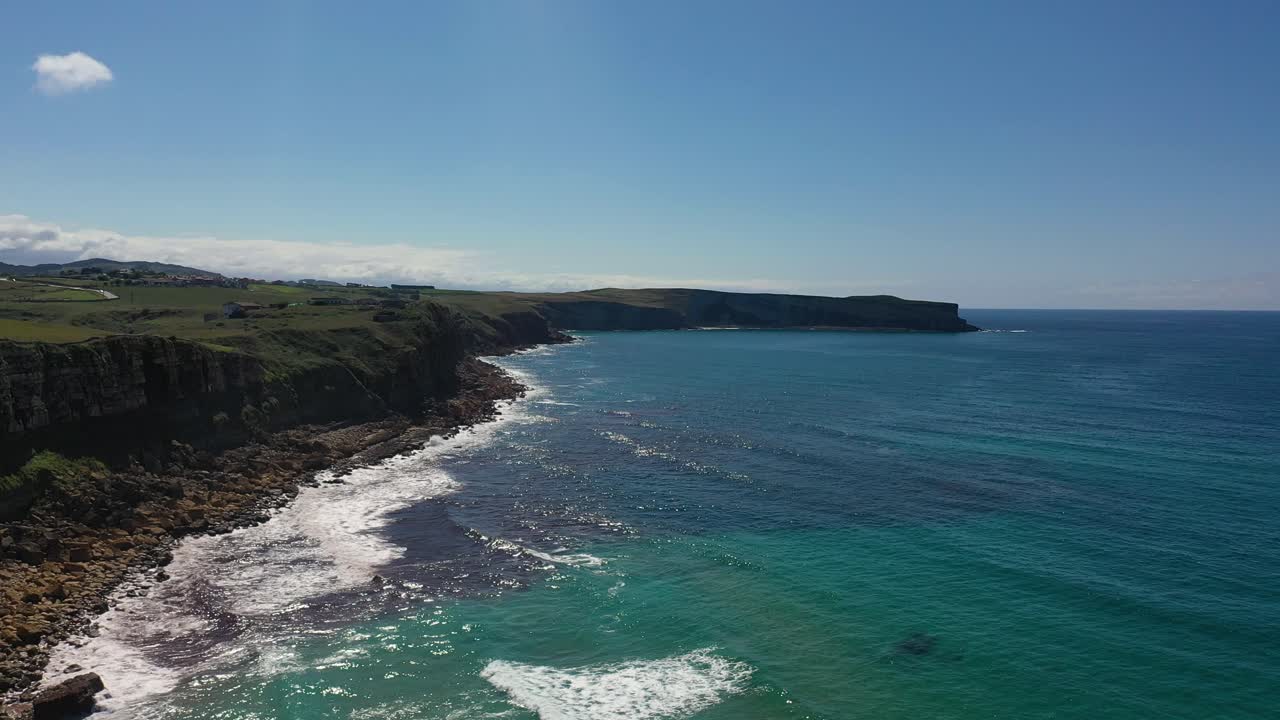 Lateral flight from the coast where you see a road and a cliff with green meadows towards the stony beach where the waves collide and the blue sea that meets a clear summer sky in Cantabria Spain
