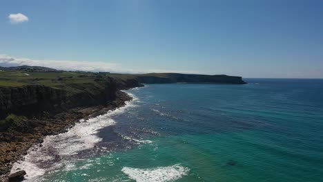 Seitlicher-Flug-Von-Der-Küste,-Wo-Sie-Eine-Straße-Und-Eine-Klippe-Mit-Grünen-Wiesen-Sehen,-In-Richtung-Des-Steinigen-Strandes,-Wo-Die-Wellen-Aufeinanderprallen-Und-Das-Blaue-Meer,-Das-Auf-Einen-Klaren-Sommerhimmel-In-Kantabrien,-Spanien-Trifft