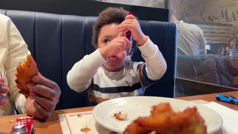 playful two-year-old black baby learns how to use a real fork eating chicken seated next to his mother inside a restaurant