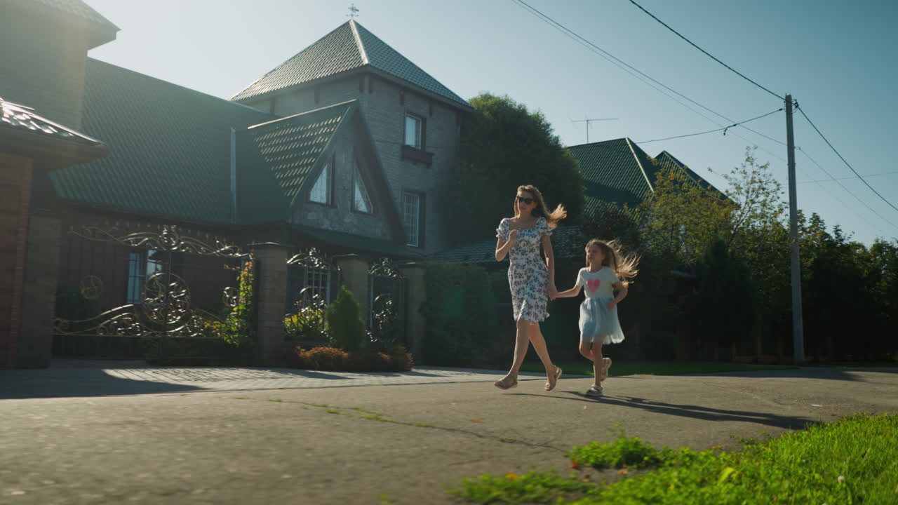 Lady in floral dress running with young girl on sunny day, holding hands while sunlight creates soft silhouette effect, casting long shadows along residential street with green trees and houses