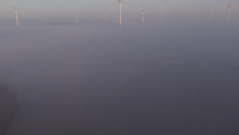 Reveal-shot-of-group-windturbines-above-the-low-fog-during-sunrise,-Aerial