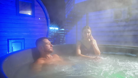 people sit in the hot tub and lots of vapor raises above them. lady in stripy swimsuit speaks on the phone.