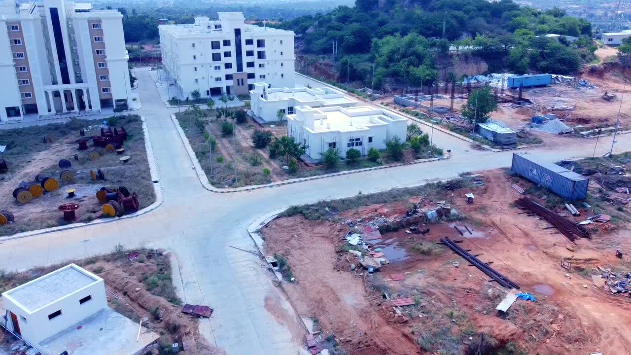 Aerial drone shot pulling back over a large construction and development site in India. The video reveals newly completed modern buildings adjacent to cleared land, raw materials, and infrastructure