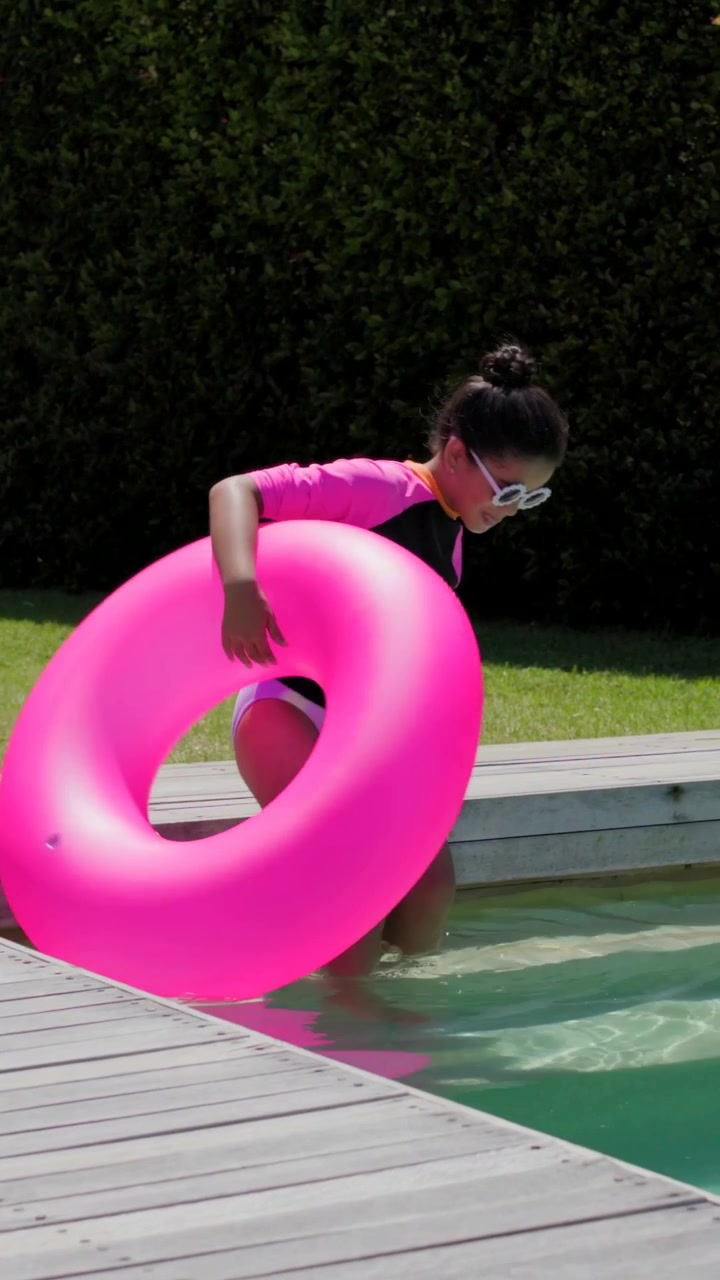Vídeo vertical: Reagindo a um braço estendendo-se, menina de rashguard deslizando em uma boia rosa, entrando na piscina de casa.