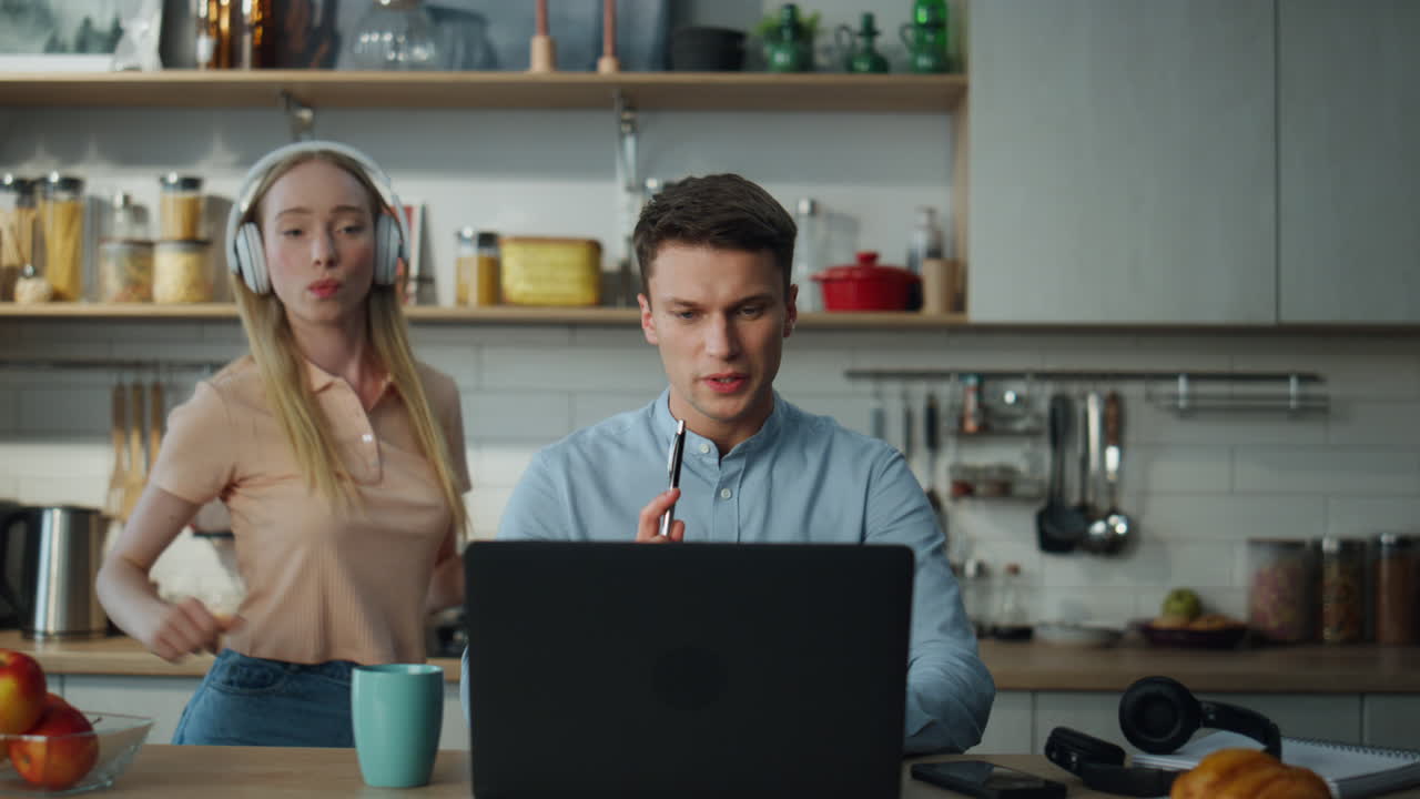 Man reporting online work results at kitchen while woman dancing on backdrop.