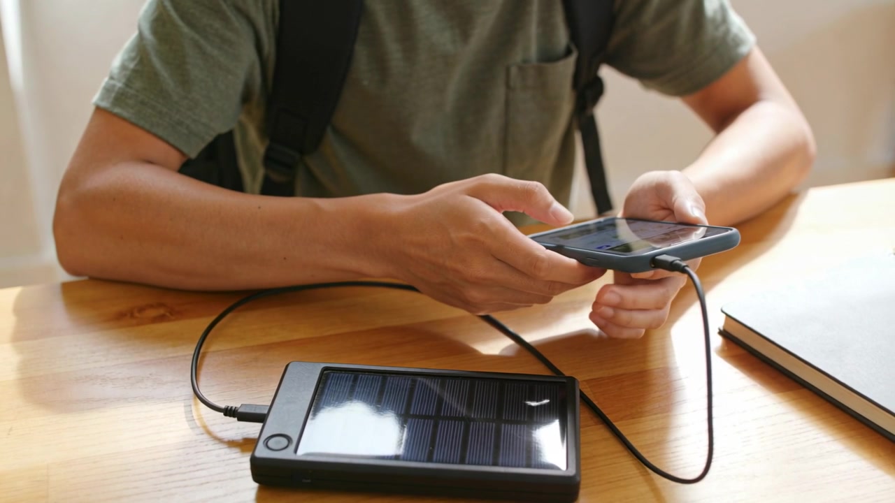 Uomo con maglietta verde che tiene e tocca un telefono con custodia blu a casa, un cavo USB avvia la ricarica di un power bank solare.