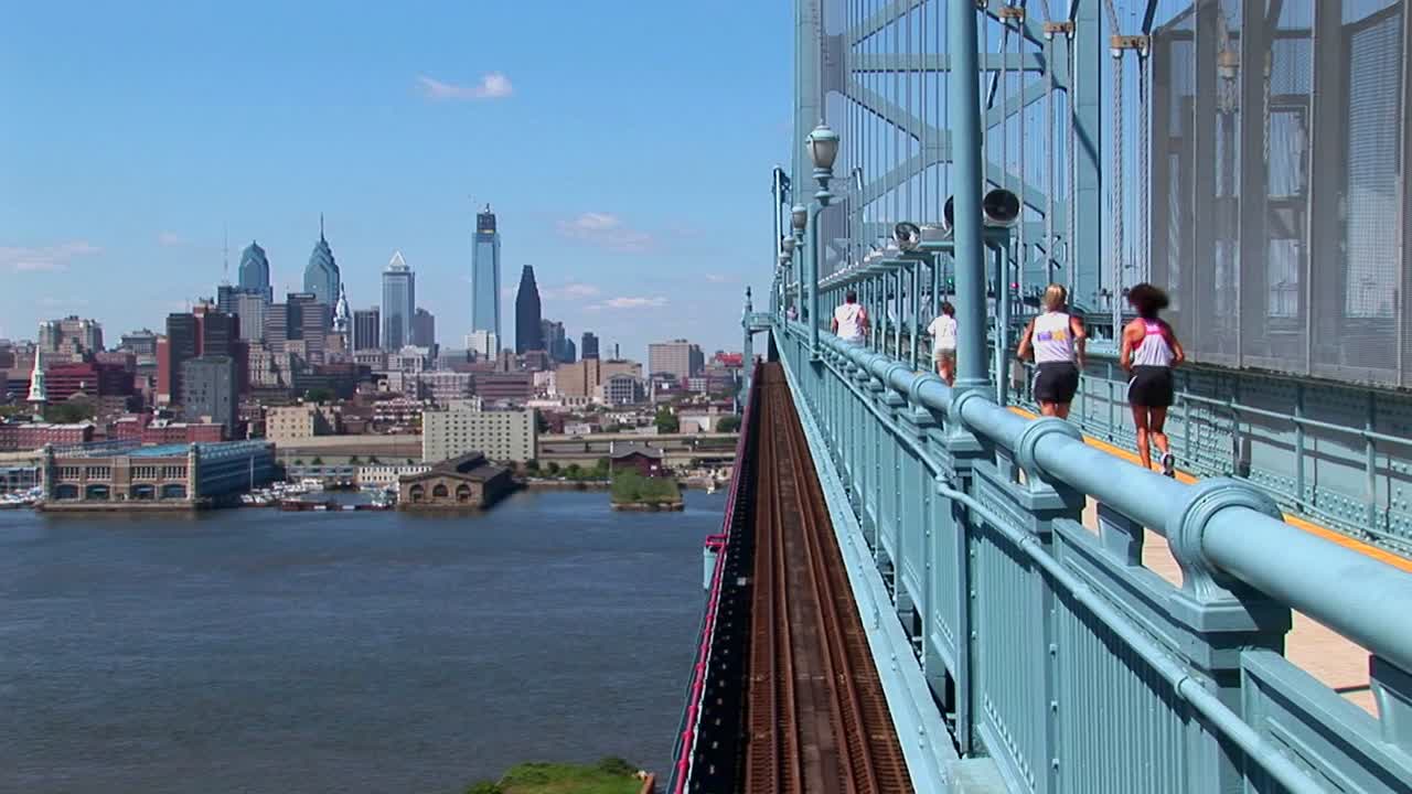 한 무리의 조깅하는 사람들이 펜실베니아 필라델피아로 이어지는 벤 프랭클린 다리를 건너고 있습니다.