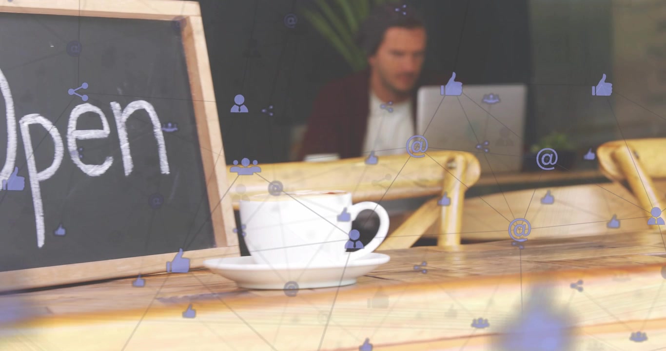Hombre escribiendo en un portátil en una cafetería creando iconos para marketing, pizarra revelando 'Abierto', taza en primer plano.