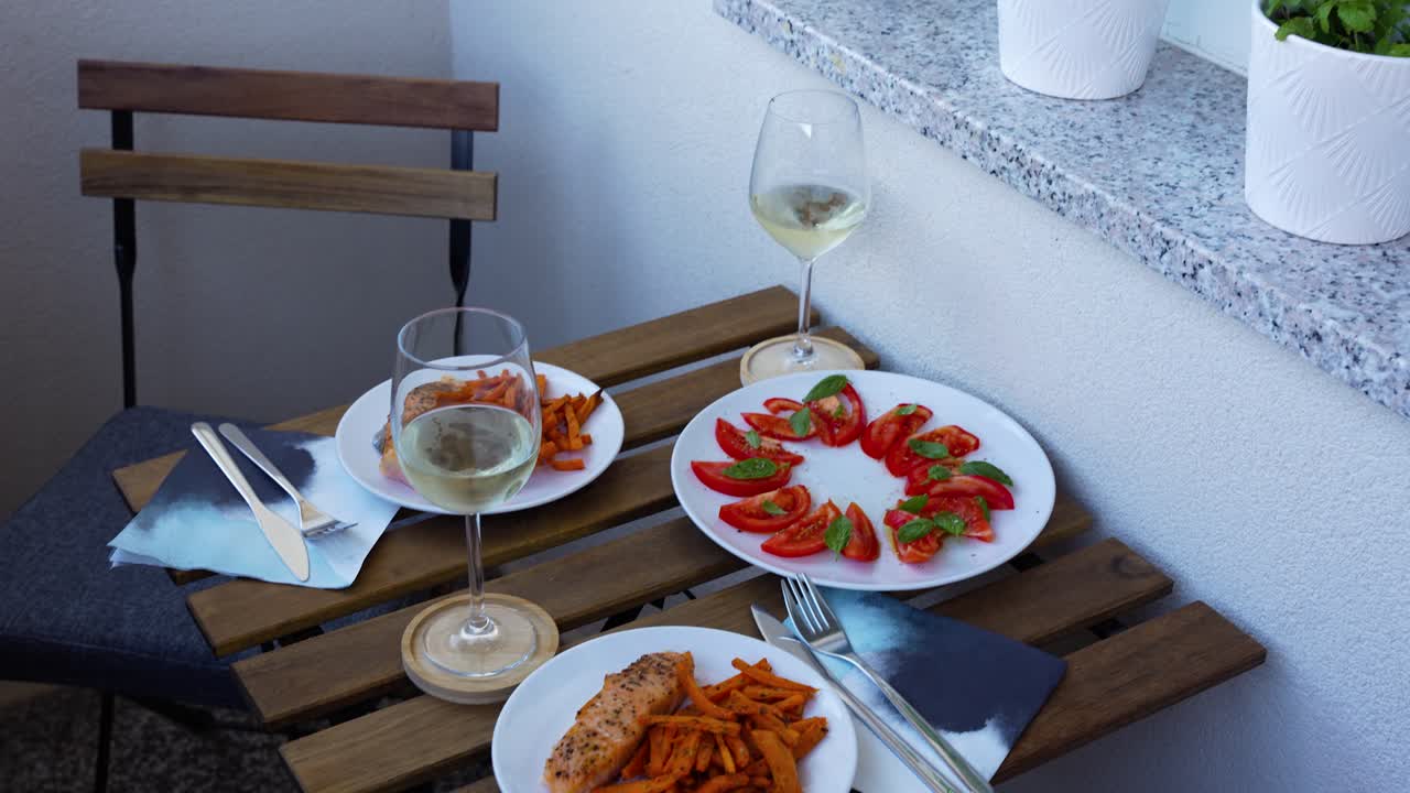 A close-up shot of a small, sunlit balcony table set for an Italian-inspired meal, featuring plates of Caprese-style tomatoes and two glasses of white wine. A hand is placing a plate onto the table