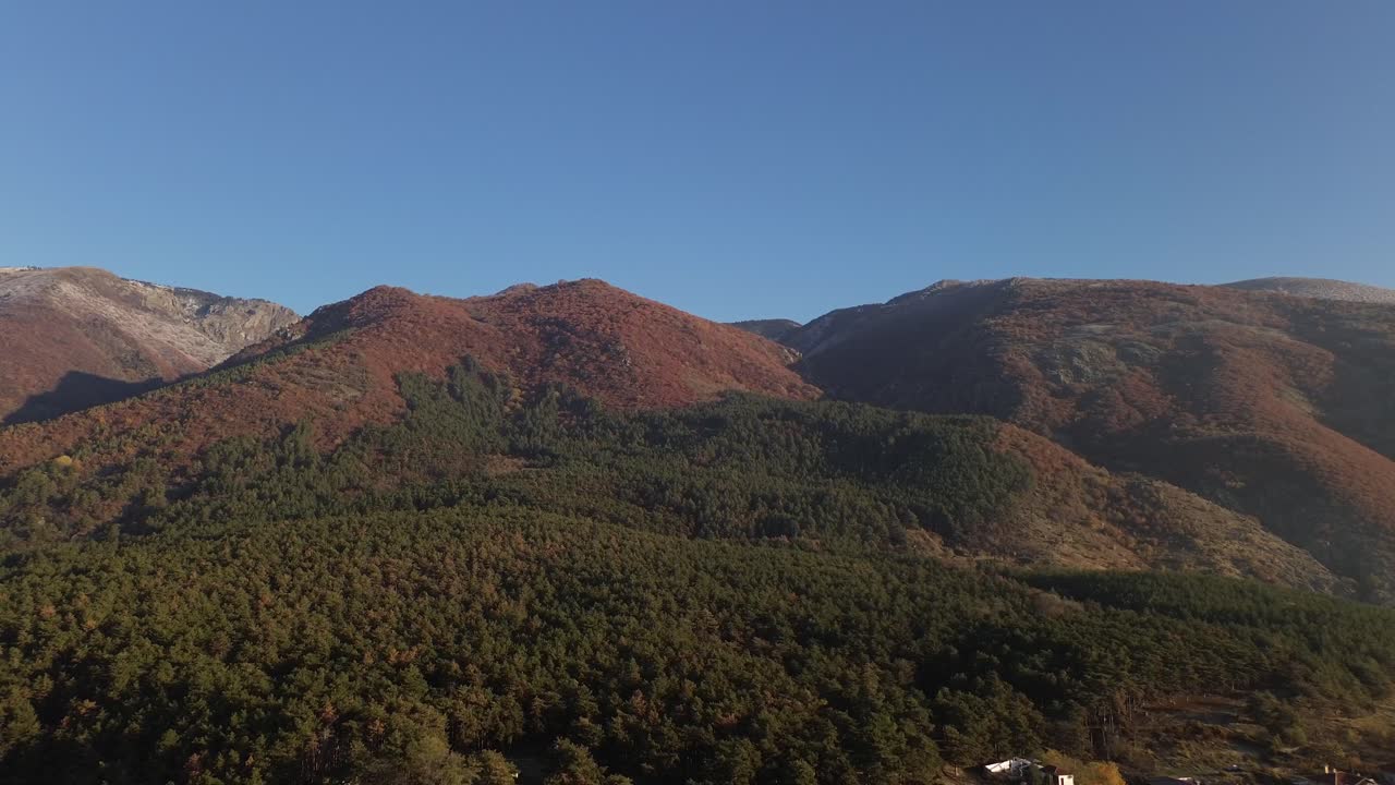 Stunning morning drone footage starting with the Balkan Mountains and a pine forest, pulling back to reveal Sopot, Bulgaria, bathed in golden hour light under a clear blue autumn sky