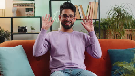 hombre indio sonriendo amistosamente a la cámara, agitando las manos gestando hola, hola, saludando en casa en el sofá