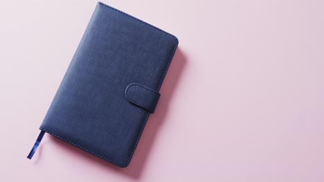 Close-up-of-closed-blue-notebook-with-copy-space-on-pink-background-in-slow-motion