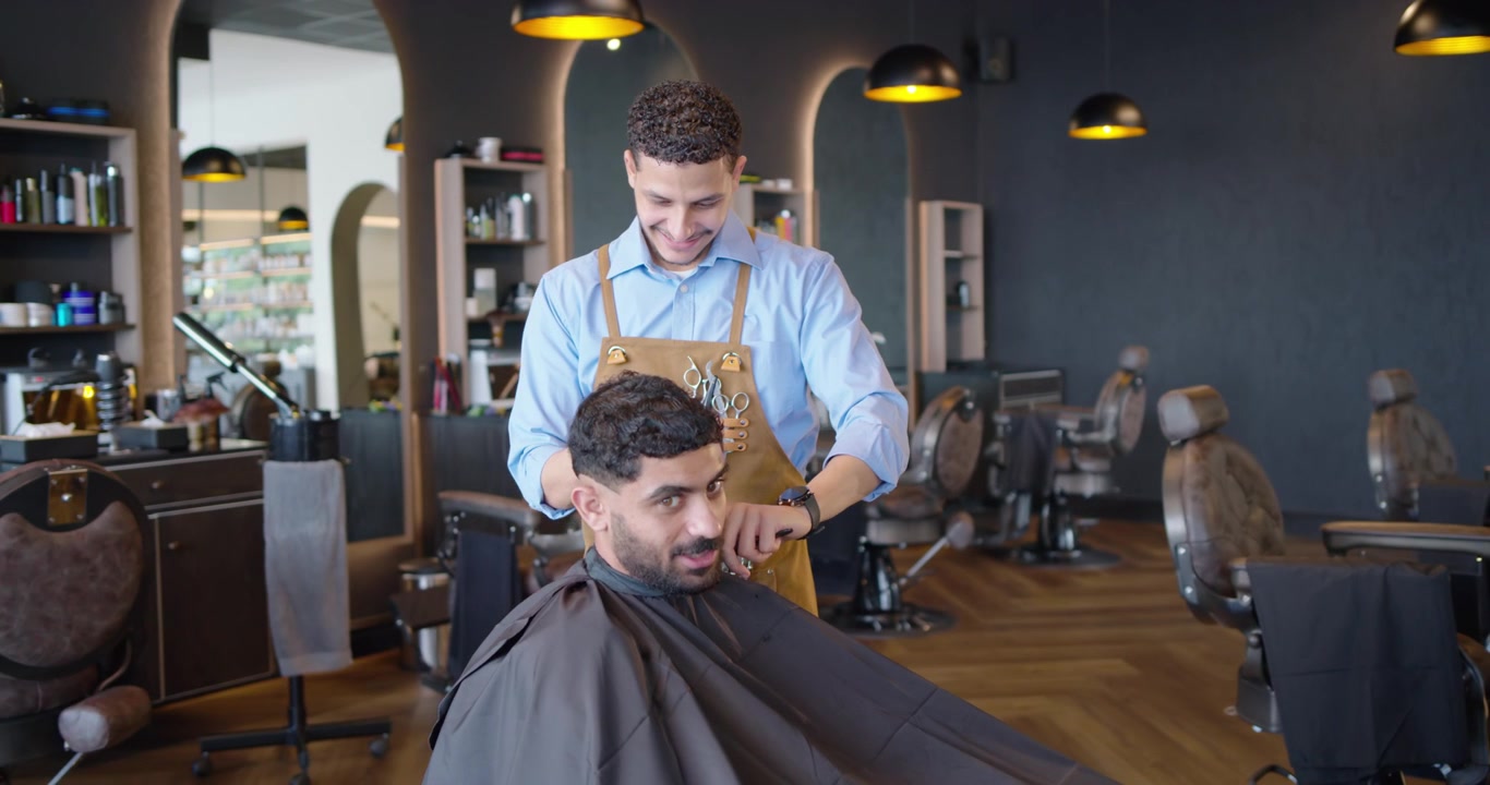 Afroamerikansk barber klipper en kunde, der sidder i en barberstol iført en sort kappe, for en pæn klipning.