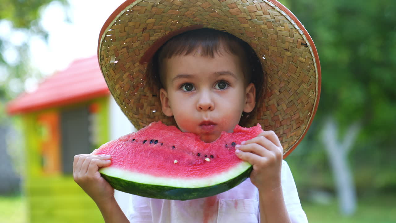 Sweet baby boy wearing a big straw hat eating tasty ripe watermelon. Healthy fruit snack outdoors in summer. Close up. Blurred backdrop.