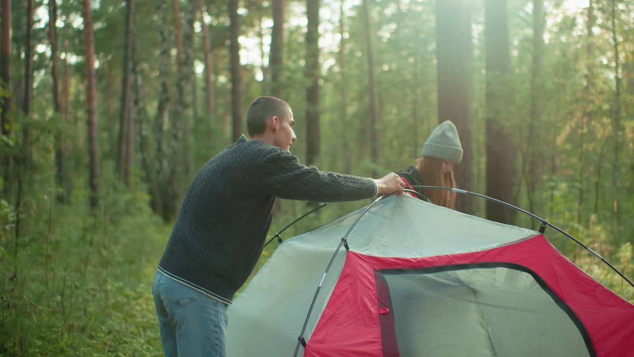 young man and woman take down camping tent in forest with warm sunlight shining through trees as girl smiles and both hold flexible poles working together in peaceful nature setting