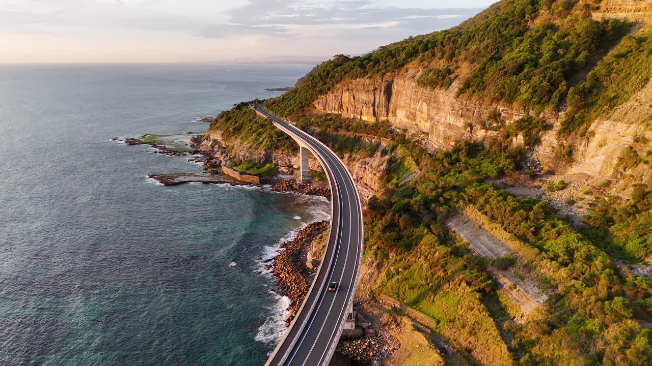 Cinematic tracking drone footage of a vehicle along the iconic Sea Cliff Bridge, showcasing stunning coastal scenery. Aerial footage golden hour.