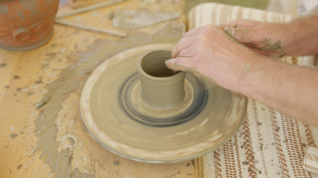 A close-up shot captures the skilled hands of a potter shaping wet clay into a pot on a spinning throwing wheel. The artisan creates handmade ceramic ware in a rustic workshop