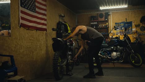 a male mechanic with a short haircut and wearing a gray tank top puts his bike on a jack in his garage workshop with tools while repairing and working with motorcycle parts
