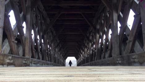 Low-wide-angle-on-pedestrian-coming-into-view-at-end-of-covered-bridge-and-walking-towards-camera,-showing-interior-wood-design