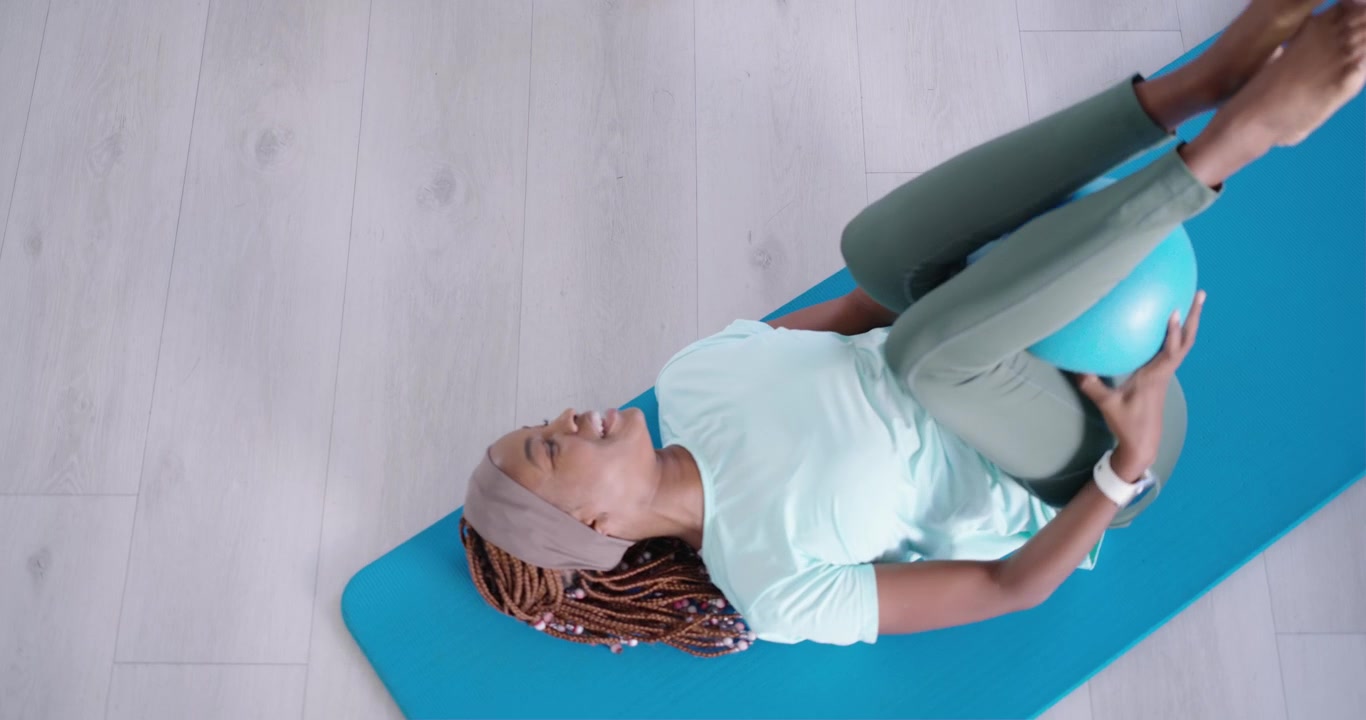 Two African women following cue on blue mats, holding blue exercise ball, lifting legs for core