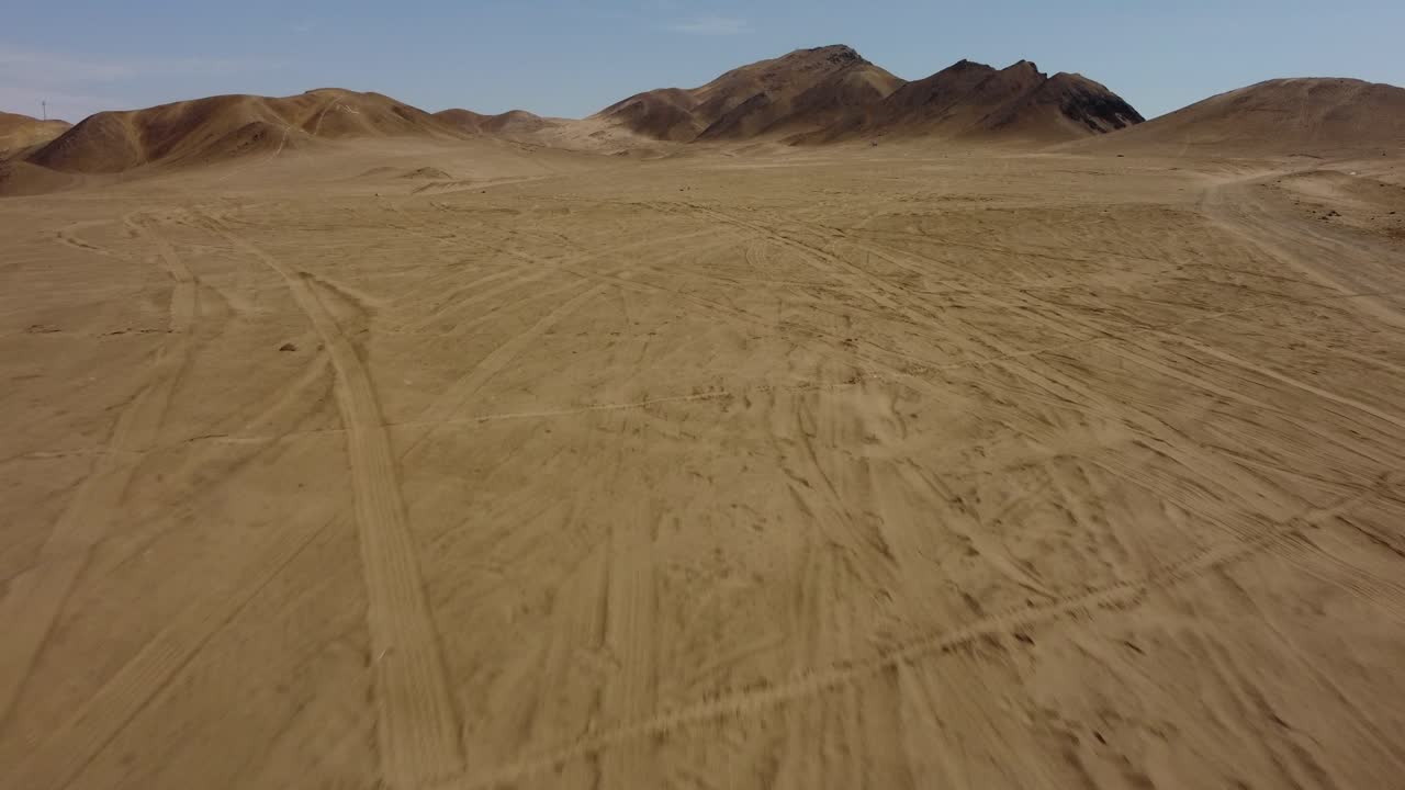 Aerial drone of a desert with dunes and mountains in the distance. Drone flies forward above tire marks in the desert then flies upwards.
