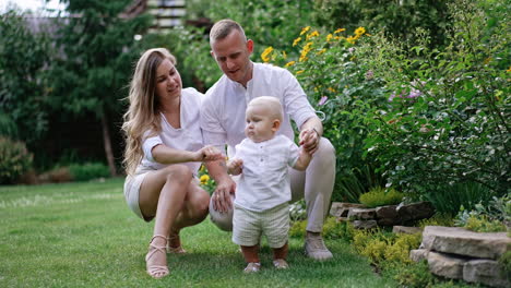 loving caucasian parents sit squatted on the grass near their cute baby. little child stands unsteadily supported by his dad.